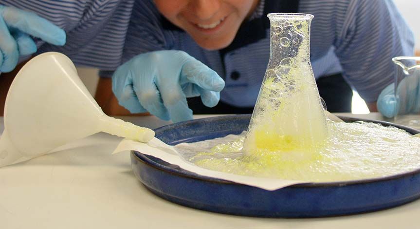 A boy's hand pointing at a beaker with yellow liquid overflowing onto a blue tray