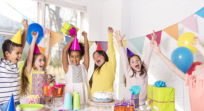 Kids around a table at a birthday party. It has decorations a cake, and candy.  Kids are having fun, their hands in the air.