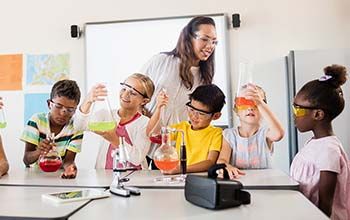 6 students looking at beakers with liquid in them.  Teacher standing over top supervising.  Also on the table is a microscope